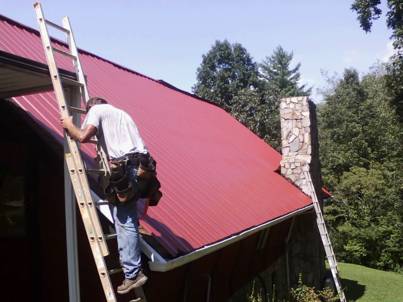 Roofer on a ladder beside a red metal roof and stone chimney — Soffit & Fascia Repair by Roofing Brothers in Bardstown